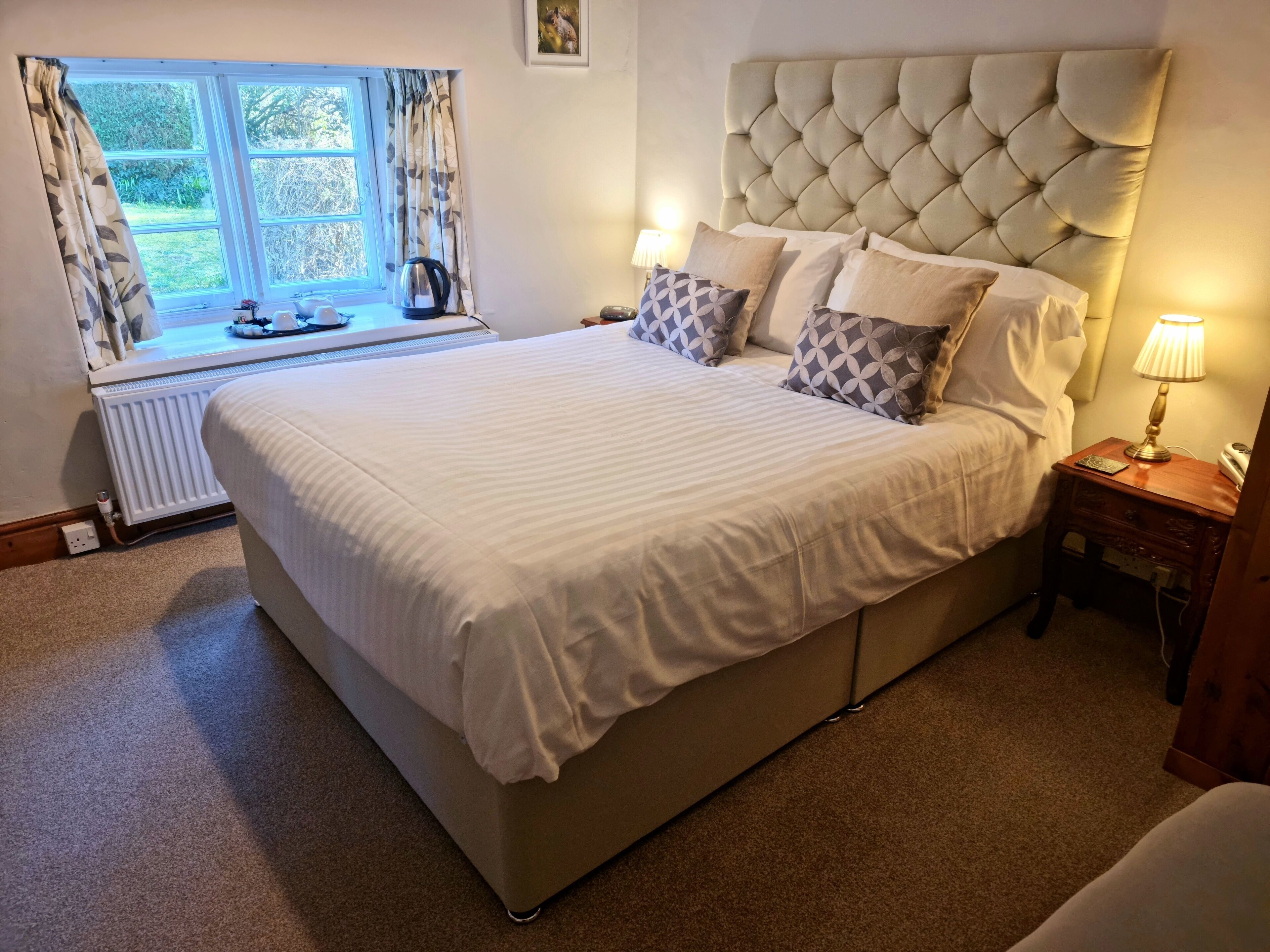 A bedroom with a large bed, a radiator, a window with curtains, and a bedside table with a lamp.