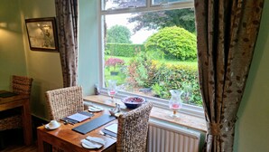 A dining area with a table set for two, a wicker chair, a radiator, and a window with curtains. - Collaven Manor Hotel (Okehampton)