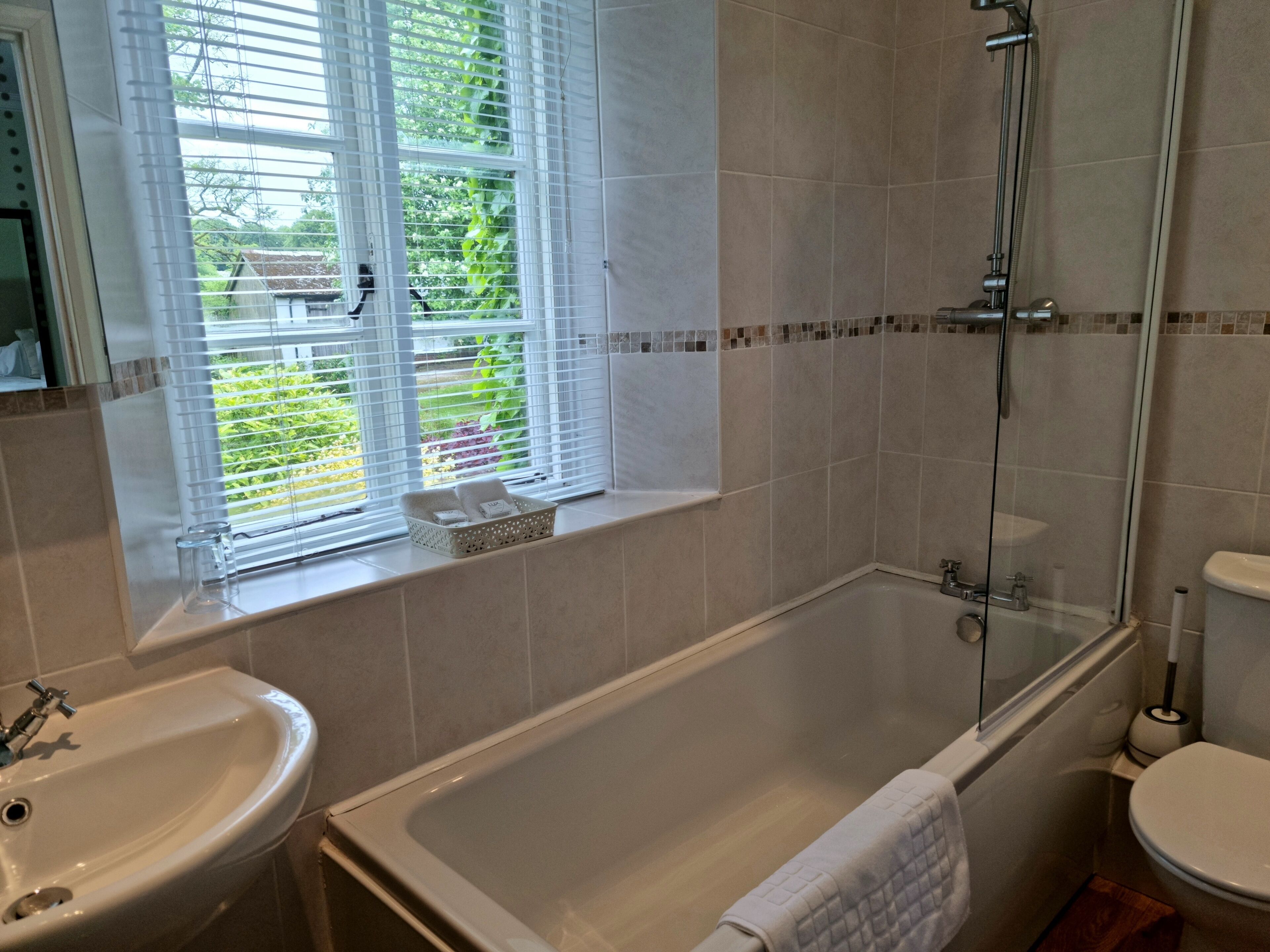 Baño con bañera, lavamanos e inodoro. Hay una ventana con persianas y vistas al exterior con vegetación.