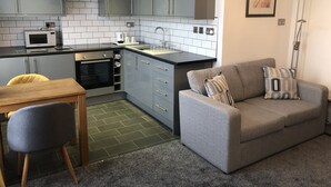 A compact kitchen with grey cabinets, a microwave, and a sink. There's a grey sofa with a cushion and a wooden table with a chair. - Edgcumbe Guest House (Plymouth)
