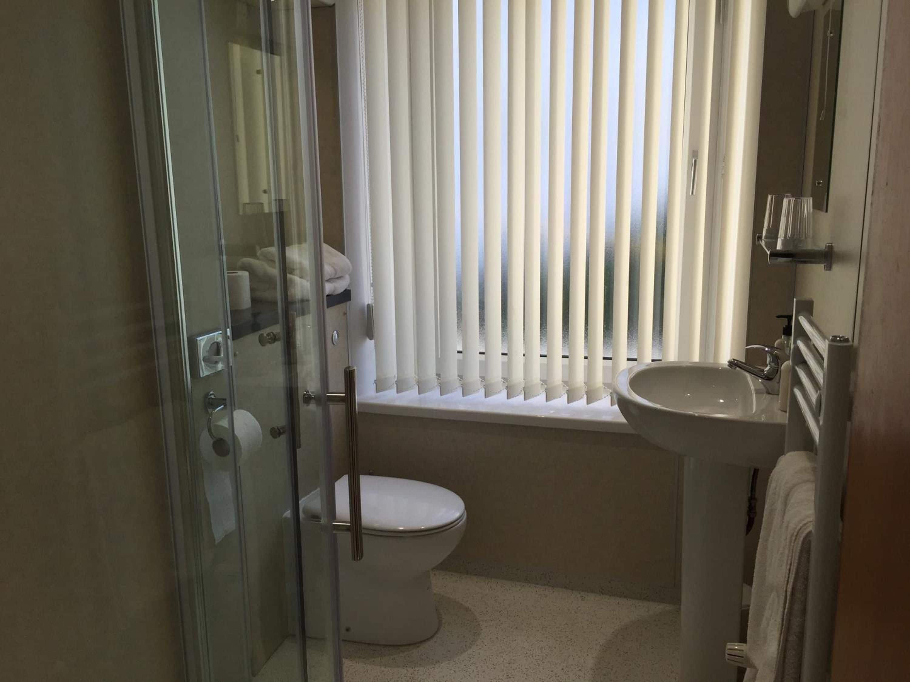 Une salle de bain avec une douche à parois de verre, des toilettes, un lavabo et une fenêtre avec des stores.