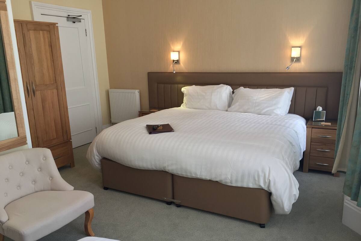 Ein Hotelzimmer mit einem großen Bett, zwei Nachttischen, einem Holzschrank, einem Sessel und einem kleinen Tisch mit einer Lampe.