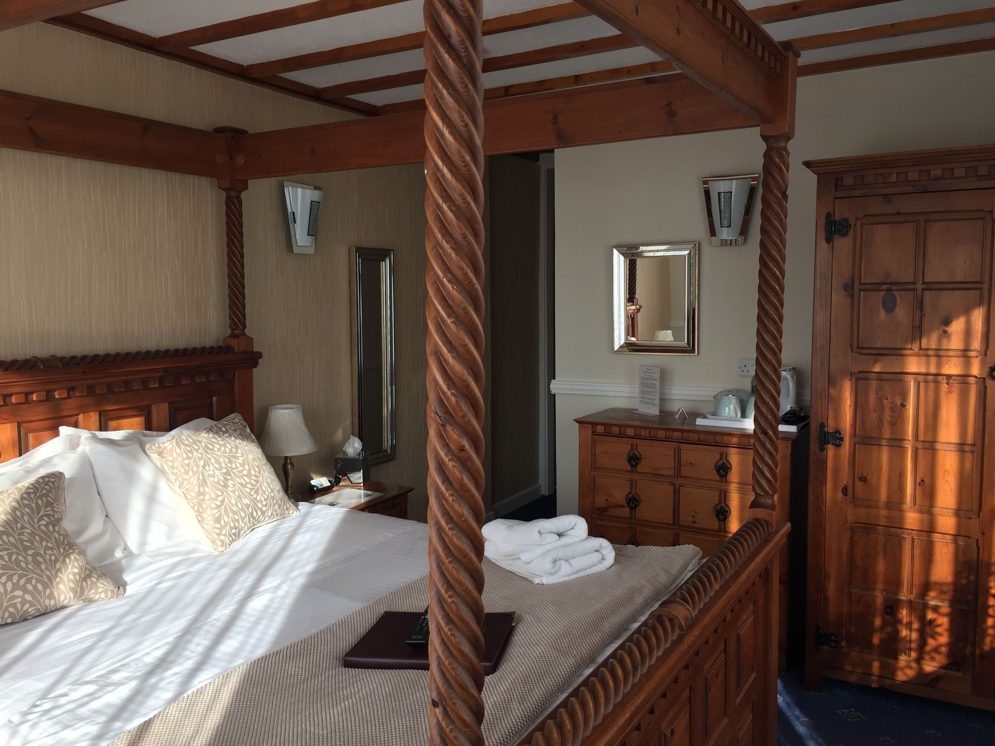 A four-poster bed with a wooden headboard, a bedside table with a lamp, a mirror, and a wooden dresser with a mirror.