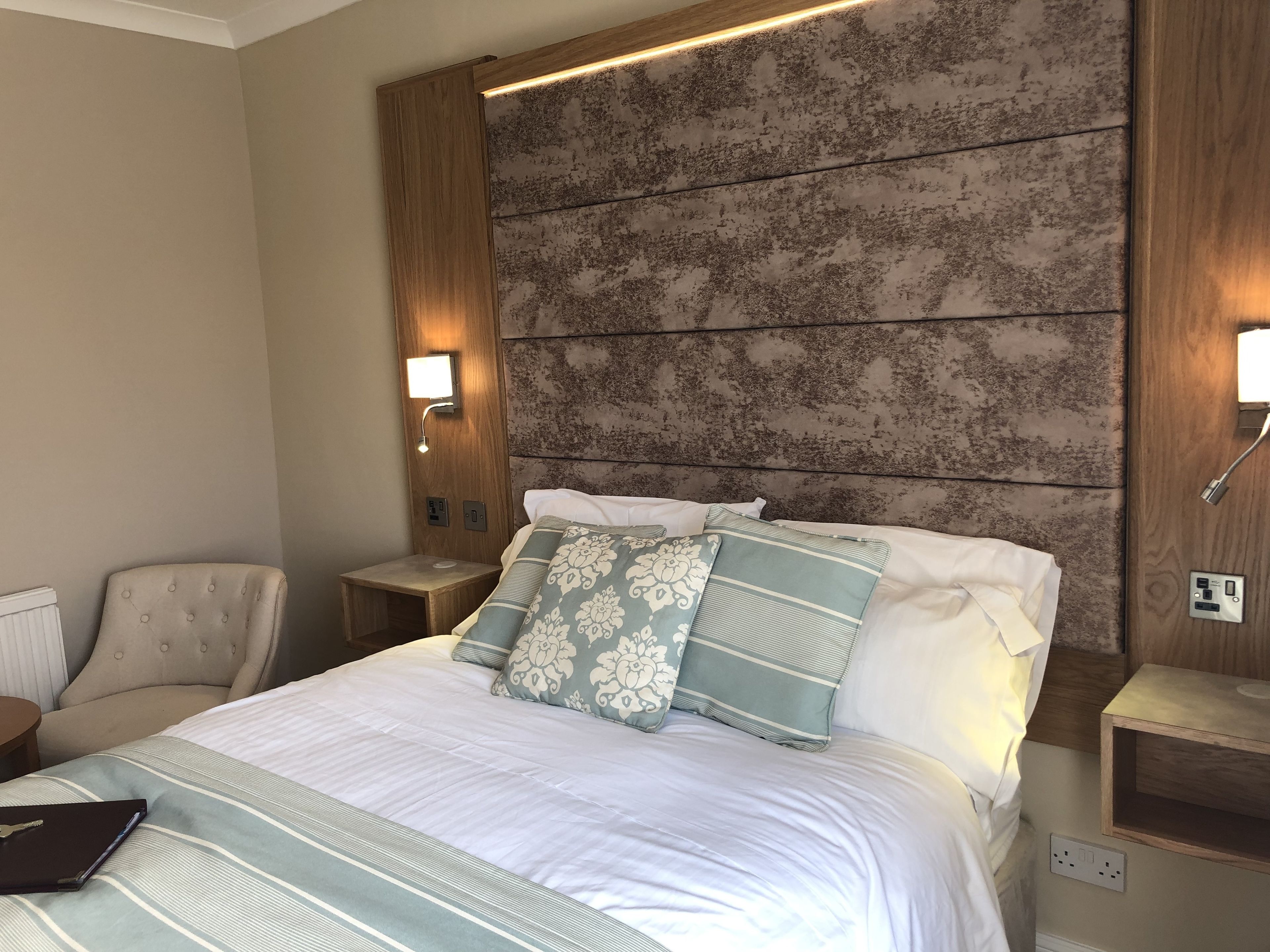 Une chambre d’hôtel avec un grand lit, deux tables de chevet, une chaise et une tête de lit dotée d’un éclairage intégré.