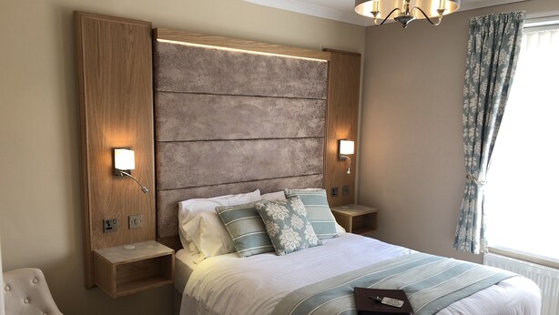 A neatly made bed with white bedding and striped pillows, a wooden headboard with built-in lighting, and a bedside table with a book. - Edgcumbe Guest House (Plymouth)