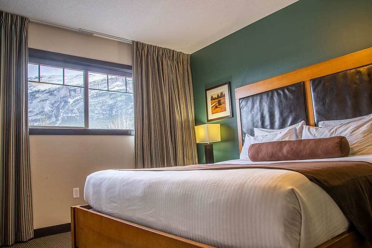 Habitación de hotel con una cama grande, una ventana con cortinas, un cuadro enmarcado en la pared y una mesita de noche con lámpara.