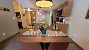A compact kitchen with wooden cabinets, a built-in oven, and a microwave. There is a dining table with two chairs and a potted plant on it.