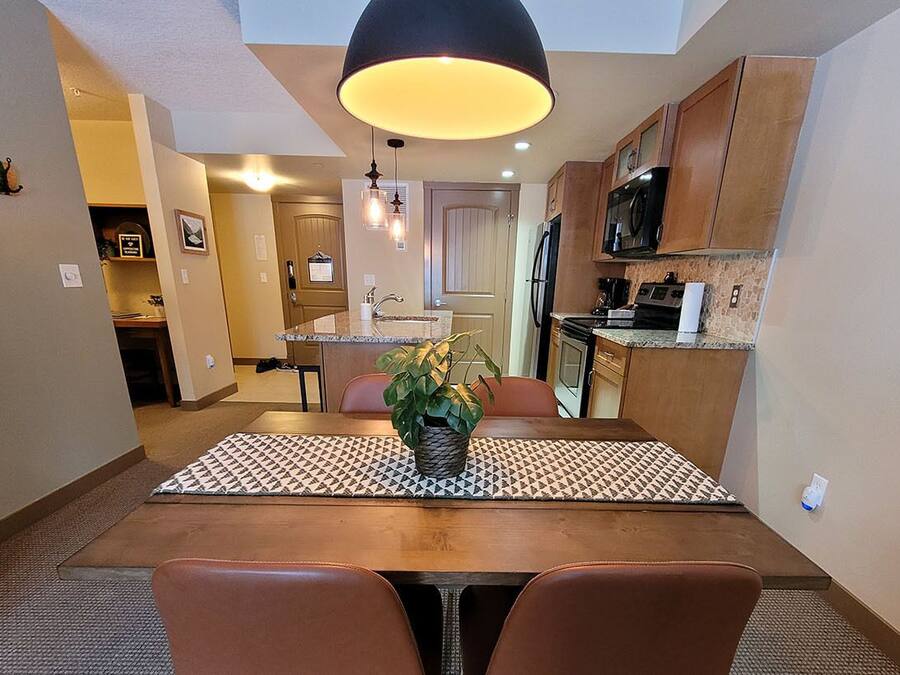 Cocina compacta con armarios de madera, horno integrado y microondas. Hay una mesa de comedor con dos sillas y una planta en maceta encima.