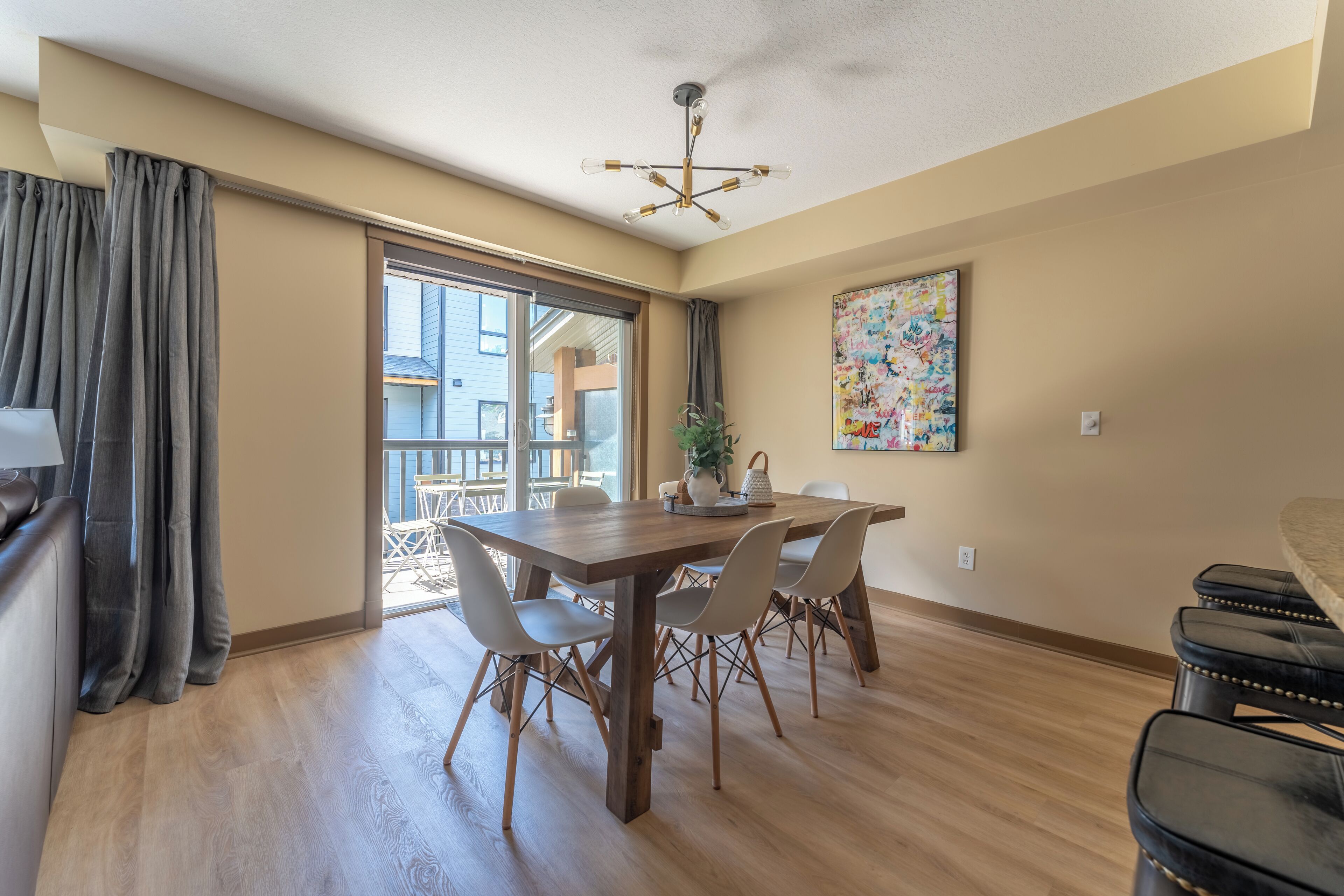 A dining area with a wooden table, six chairs, a chandelier, and a large window with curtains.
