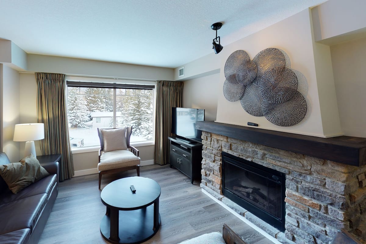 Una sala de estar con chimenea, un sofá, un televisor y vistas a árboles cubiertos de nieve.