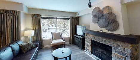 Una sala de estar con chimenea, un sofá, un televisor y vistas a árboles cubiertos de nieve.