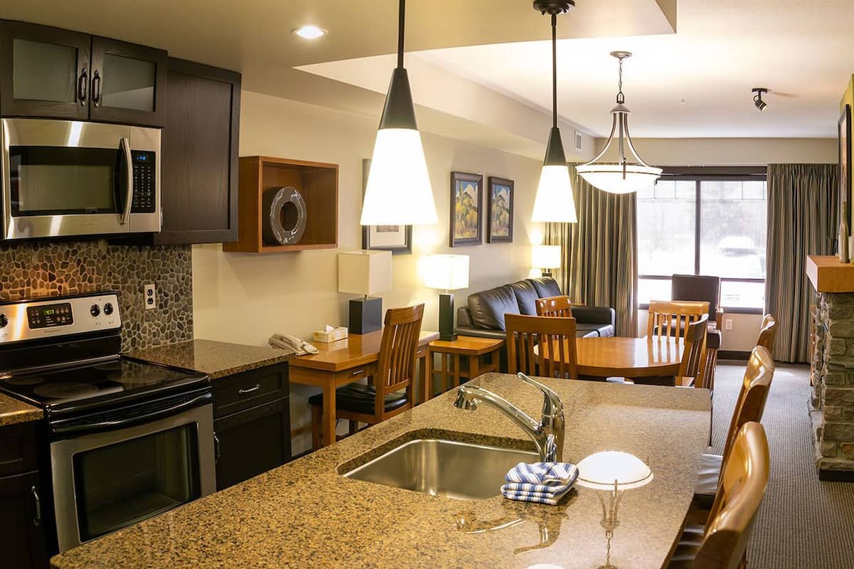 Una cocina moderna con encimeras de granito, electrodomésticos de acero inoxidable y una isla central con pileta. Se observan sillas de comedor de madera y una mesa, además de una chimenea de piedra.