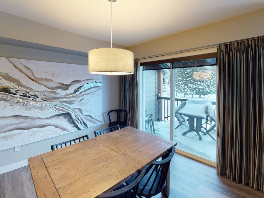 Een eethoek met een houten tafel en stoelen, een groot abstract schilderij aan de muur, en een uitzicht op een besneeuwde patio door een schuifdeur.