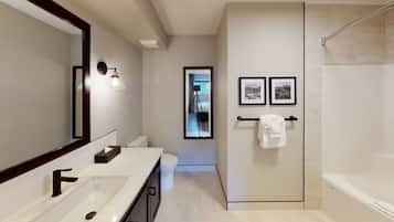 A modern bathroom with a large mirror, two sinks, a toilet, and a walk-in shower. There are framed pictures on the wall and a towel hanging on a rack.