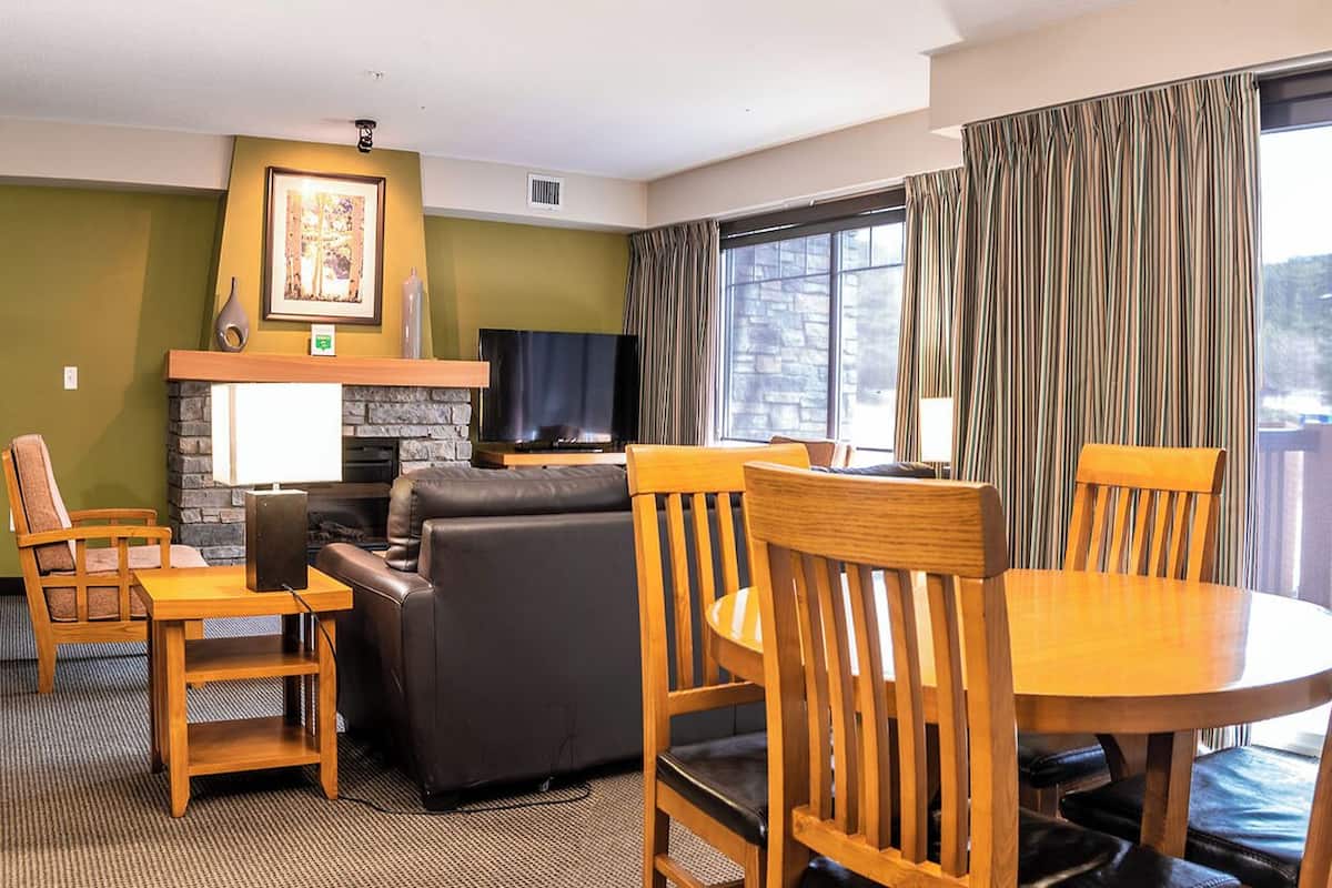 Habitación de hotel con chimenea, televisor de pantalla plana, sofá, mesa de comedor con sillas y un gran ventanal con cortinas.