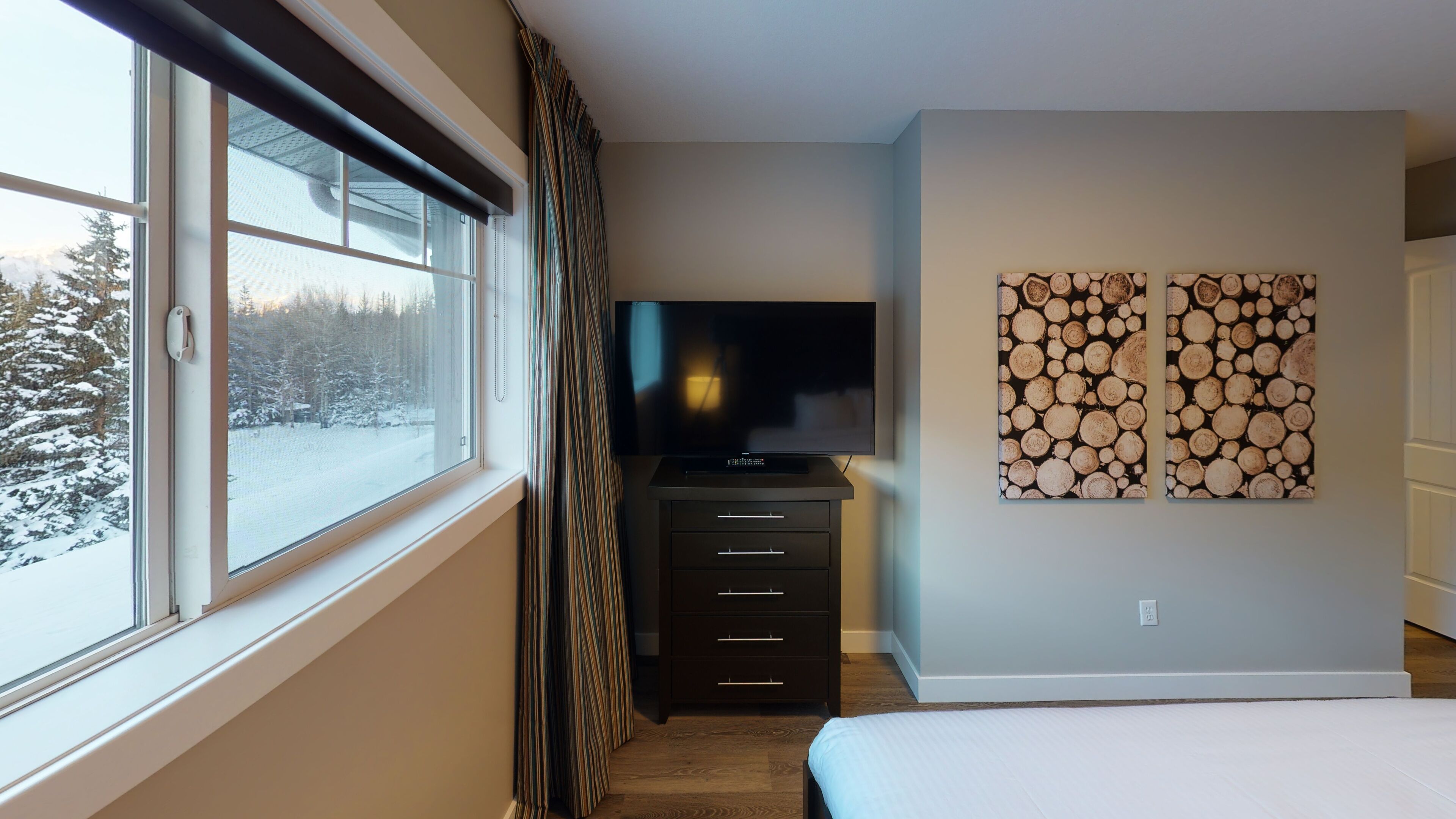 A bedroom with a large window, a flat-screen TV, a dark dresser, and a wall-mounted artwork of logs.