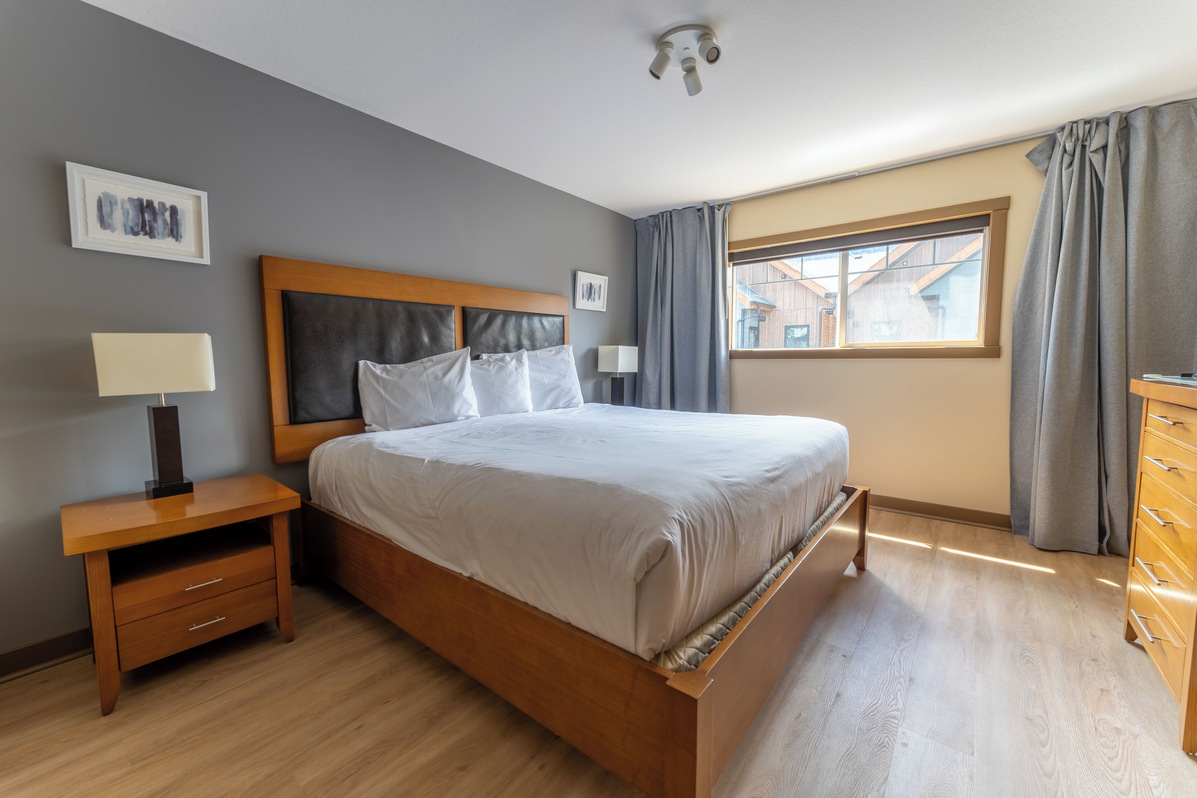 A bedroom with a wooden bed, a nightstand with a lamp, a dresser, and a window with curtains.