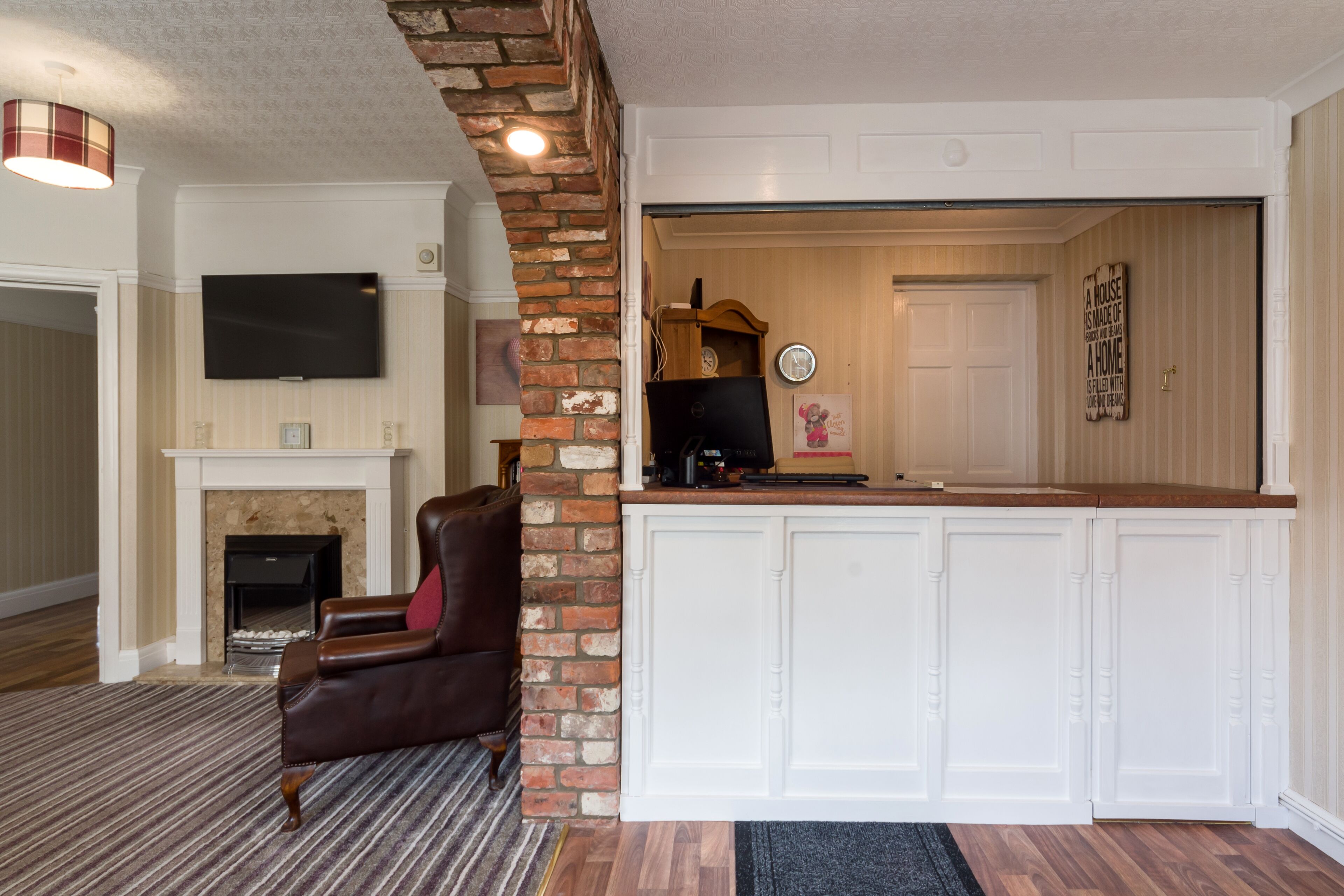 A living room with a fireplace, a leather armchair, a flat-screen TV, and a kitchen area with a brick wall and white cabinets.