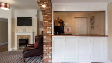 A living room with a fireplace, a leather armchair, a flat-screen TV, and a kitchen area with a brick wall and white cabinets.