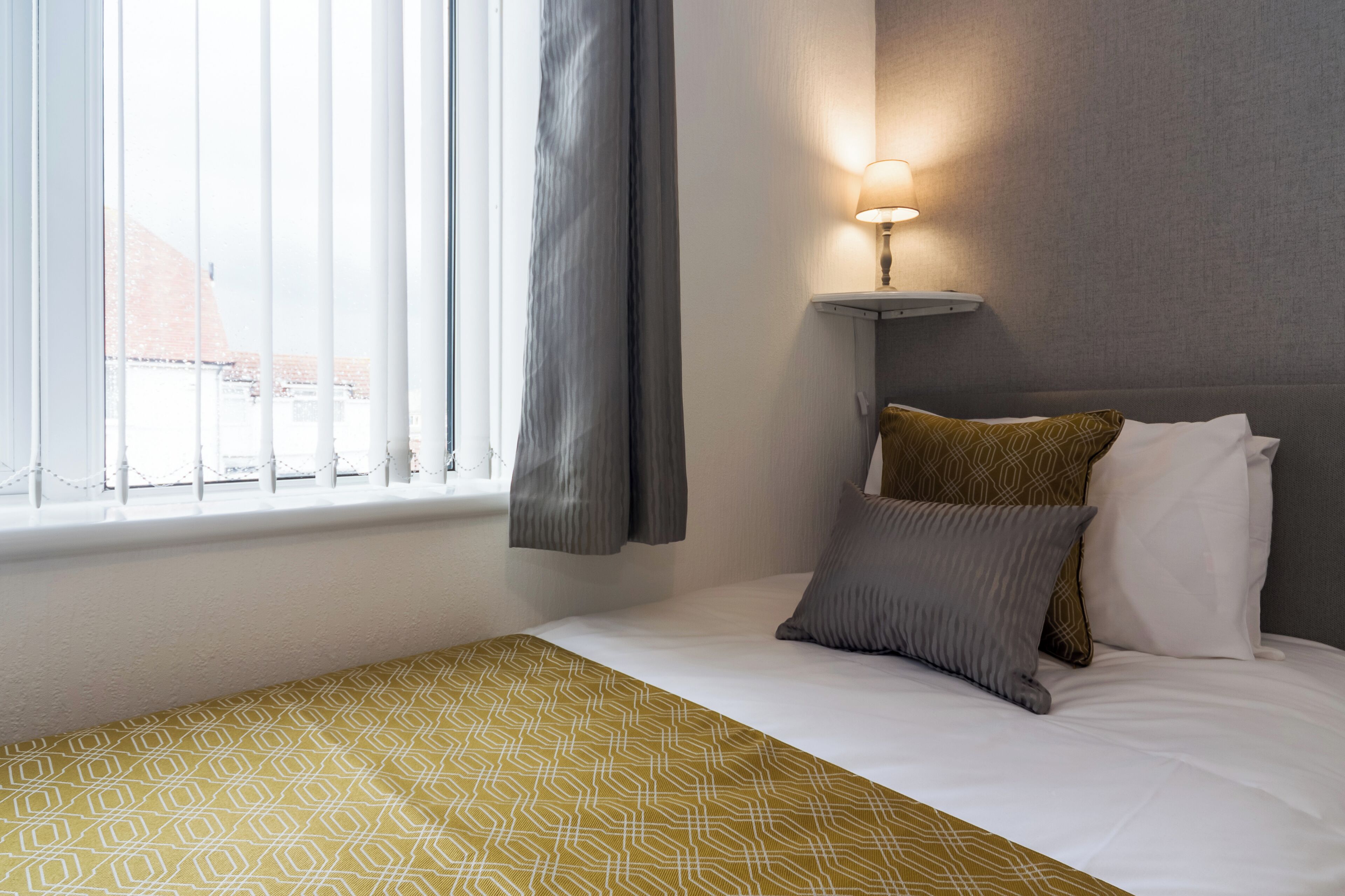 A hotel room with a bed, a bedside lamp, a window with curtains, and a patterned throw blanket.