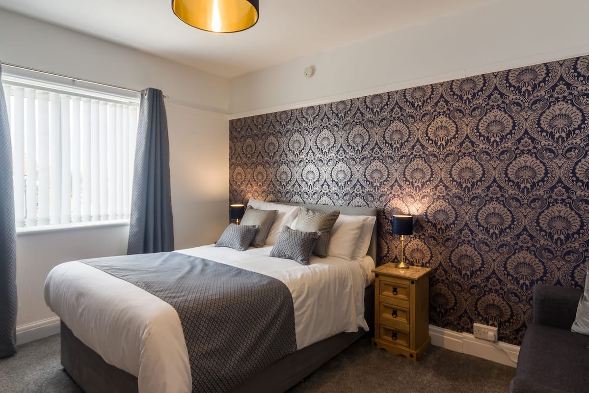 A bedroom with a large bed, bedside tables, a patterned wallpaper, and a window with curtains.