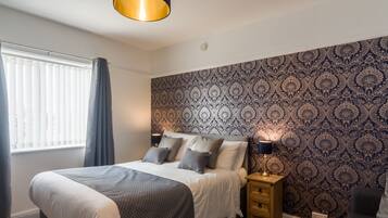A bedroom with a large bed, bedside tables, a patterned wallpaper, and a window with curtains.