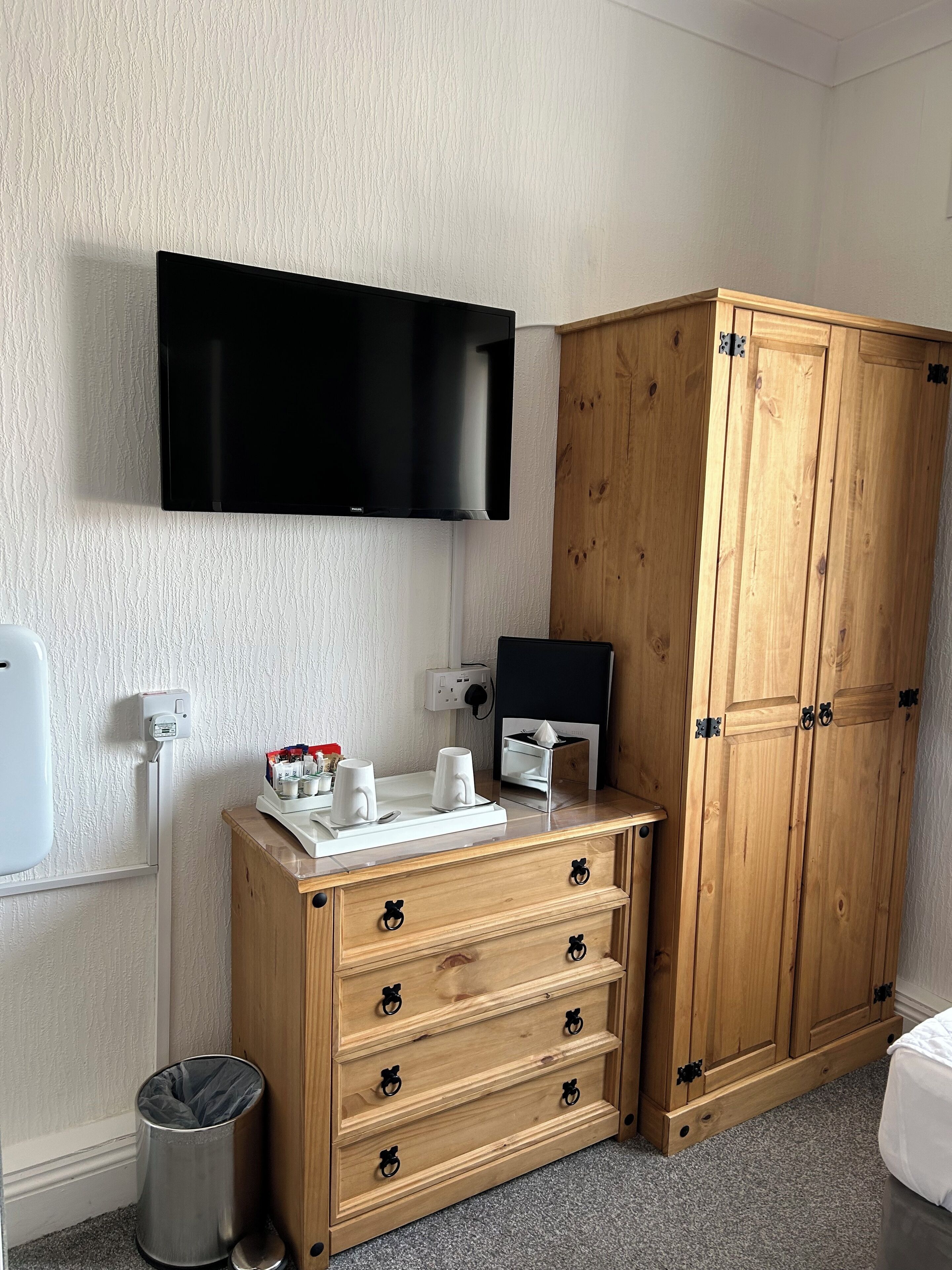 A hotel room with a wooden wardrobe, a flat-screen TV mounted on the wall, a small table with cups and a tissue box, and a trash bin.