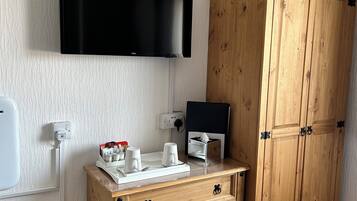 A hotel room with a wooden wardrobe, a flat-screen TV mounted on the wall, a small table with cups and a tissue box, and a trash bin.