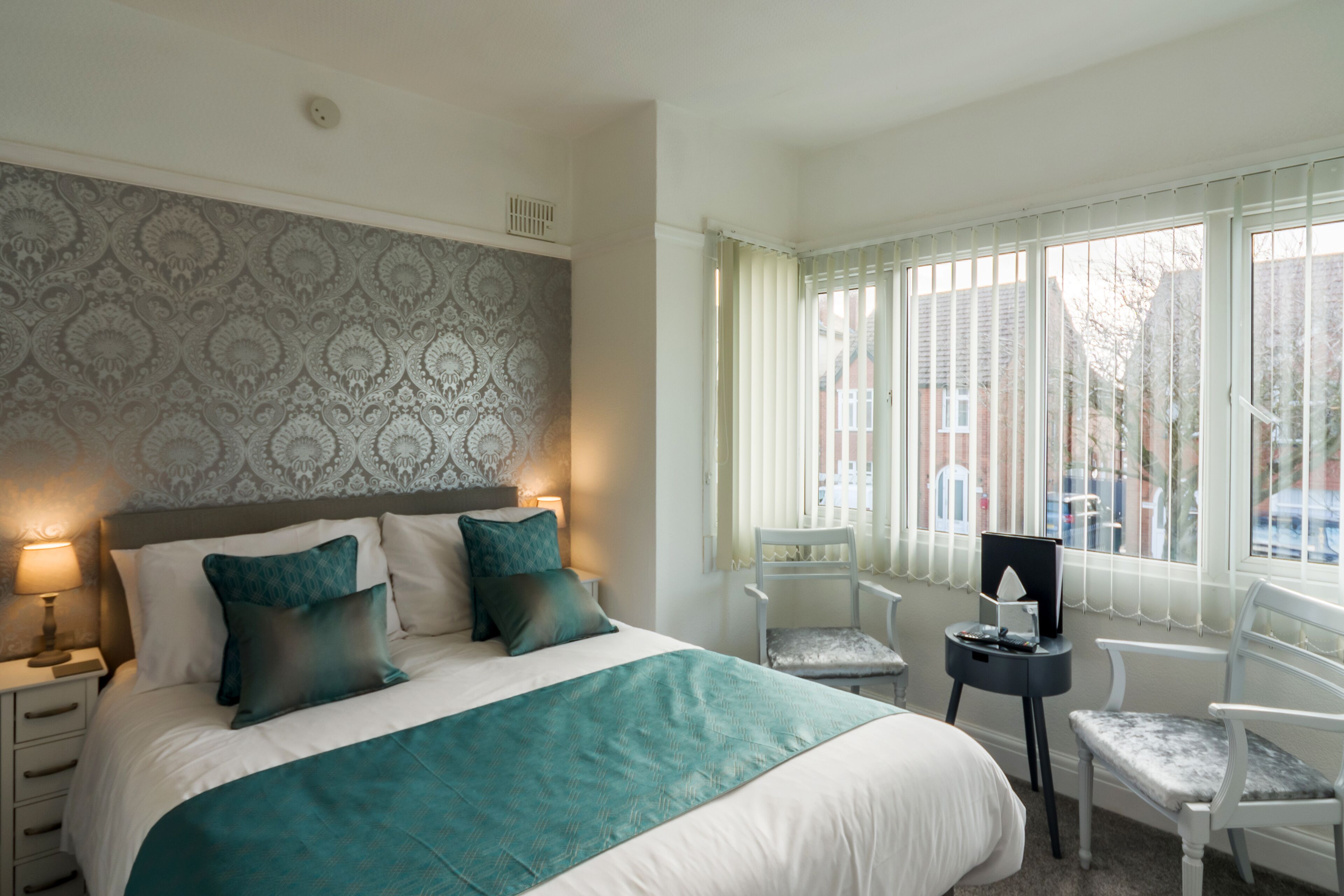 A neatly arranged hotel room with a large bed, a bedside table with a lamp, a chair, and a window with blinds.