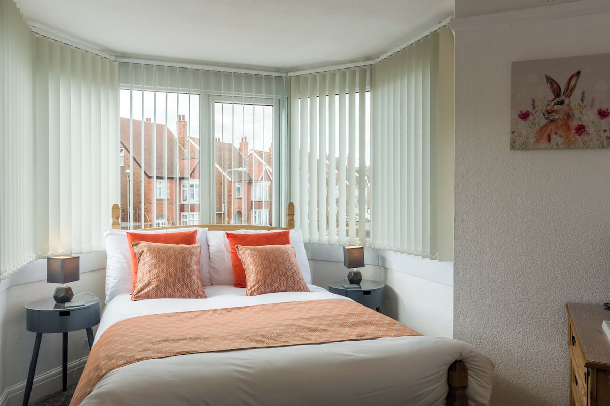 A bedroom with a large bed, two bedside tables, a nightstand, and a window with blinds.