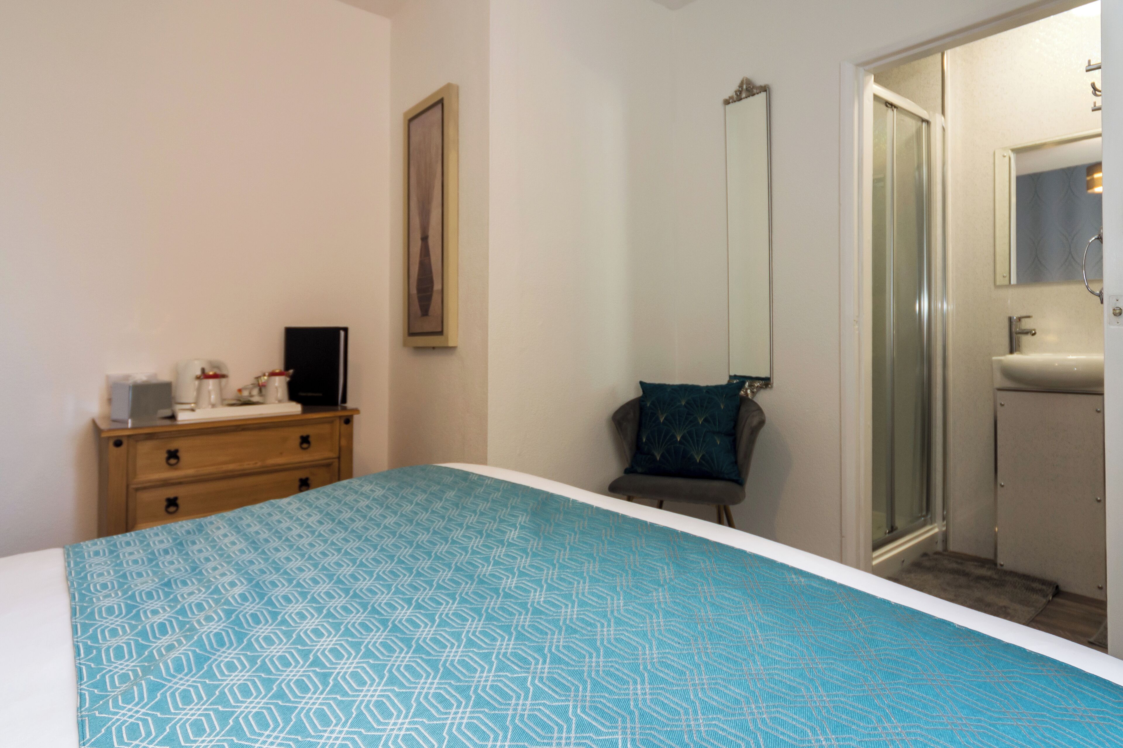 A bedroom with a bed, a dresser, a chair, a shower, and a bathroom with a sink and mirror.