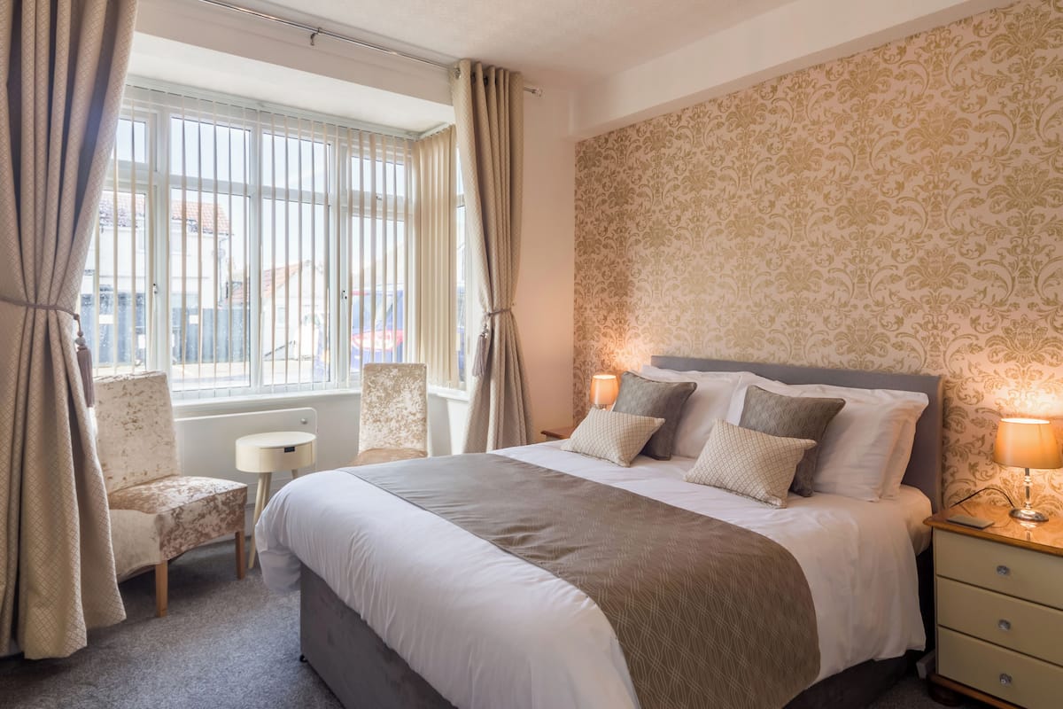 A hotel room with a large bed, a chair, a small table, and a window with curtains.