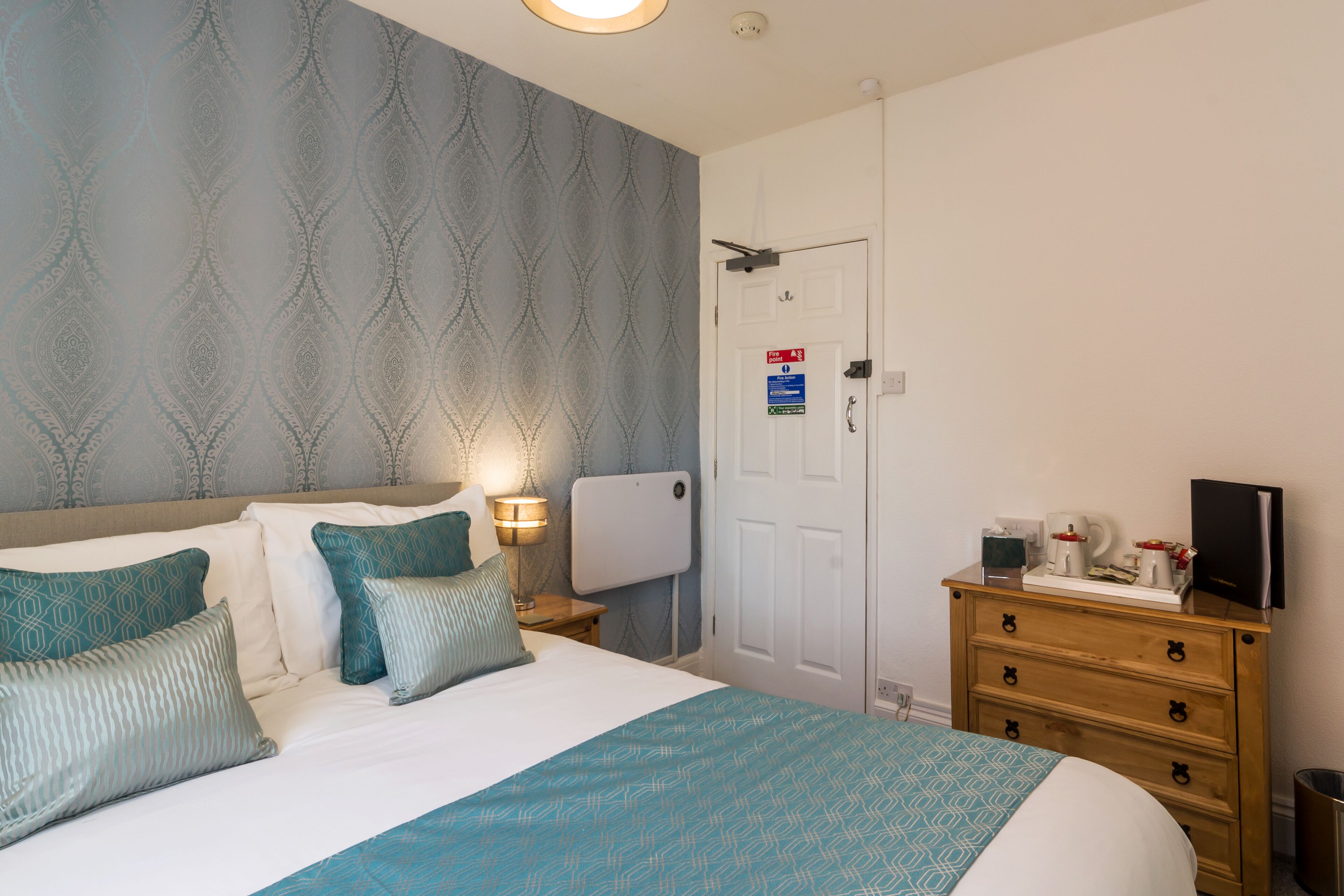 A neatly arranged hotel room with a bed, bedside table, lamp, dresser, and a door with a sign.