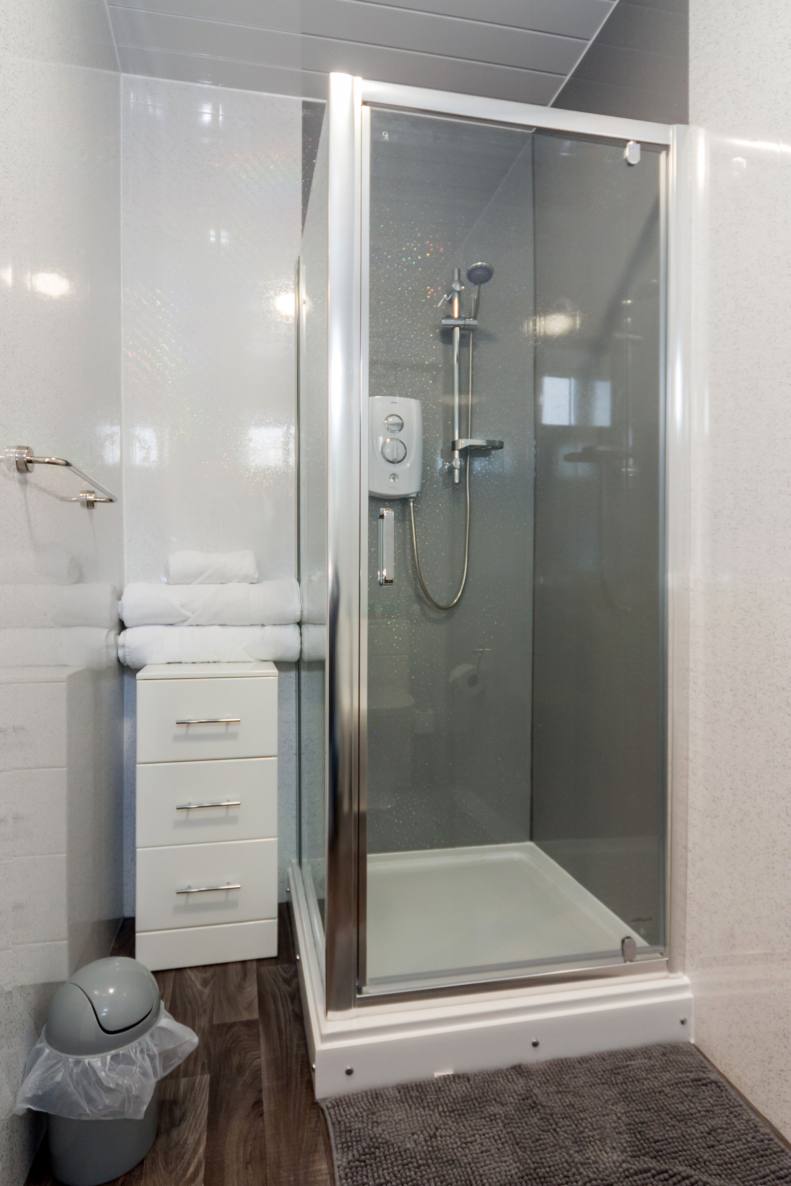A glass-enclosed shower with a control panel, a white storage unit with drawers, and a gray trash bin.