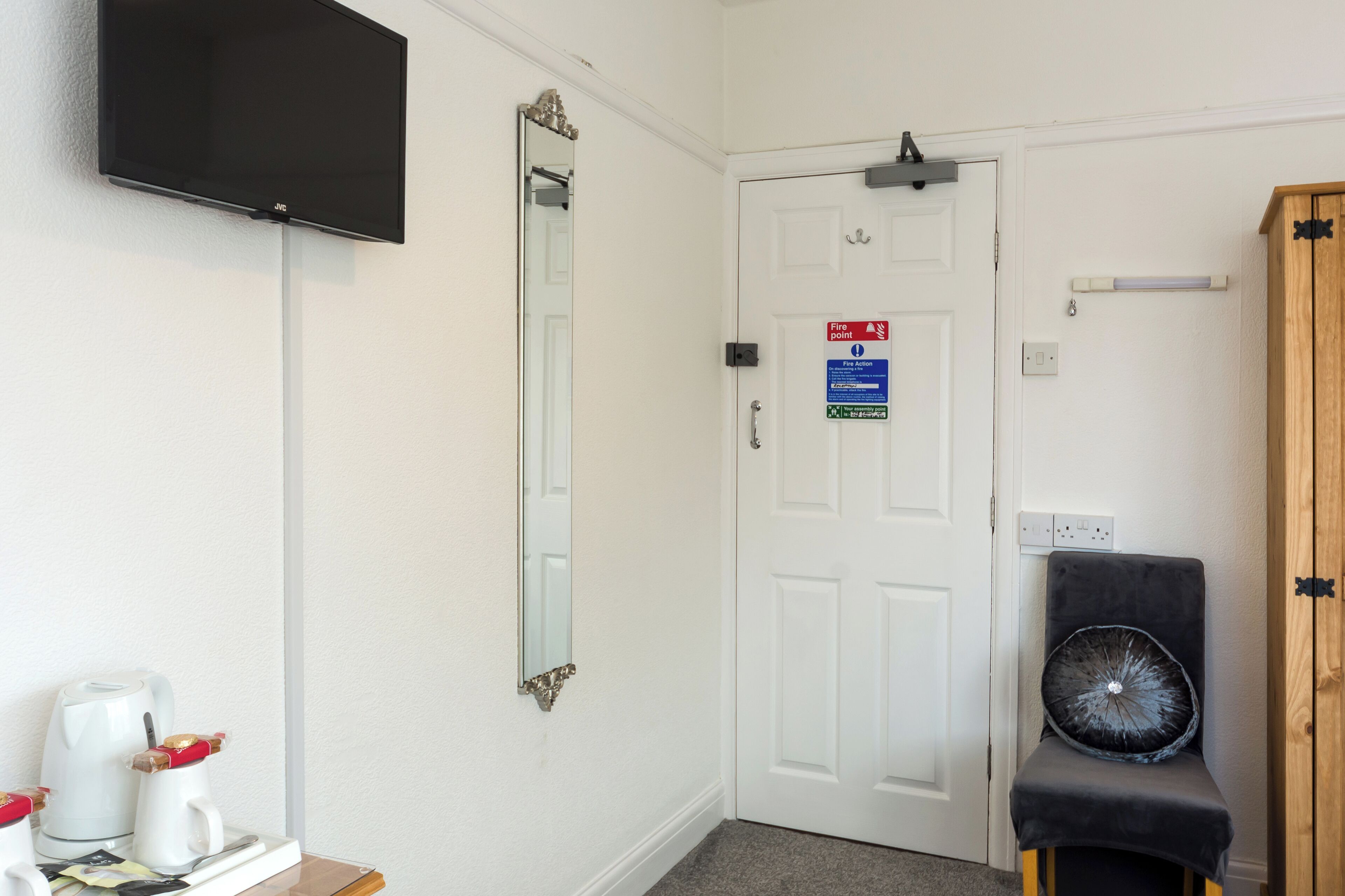 A hotel room with a flat-screen TV, a mirror, a door with a fire point sign, a small table with a kettle and cups, and a chair with a decorative cushion.