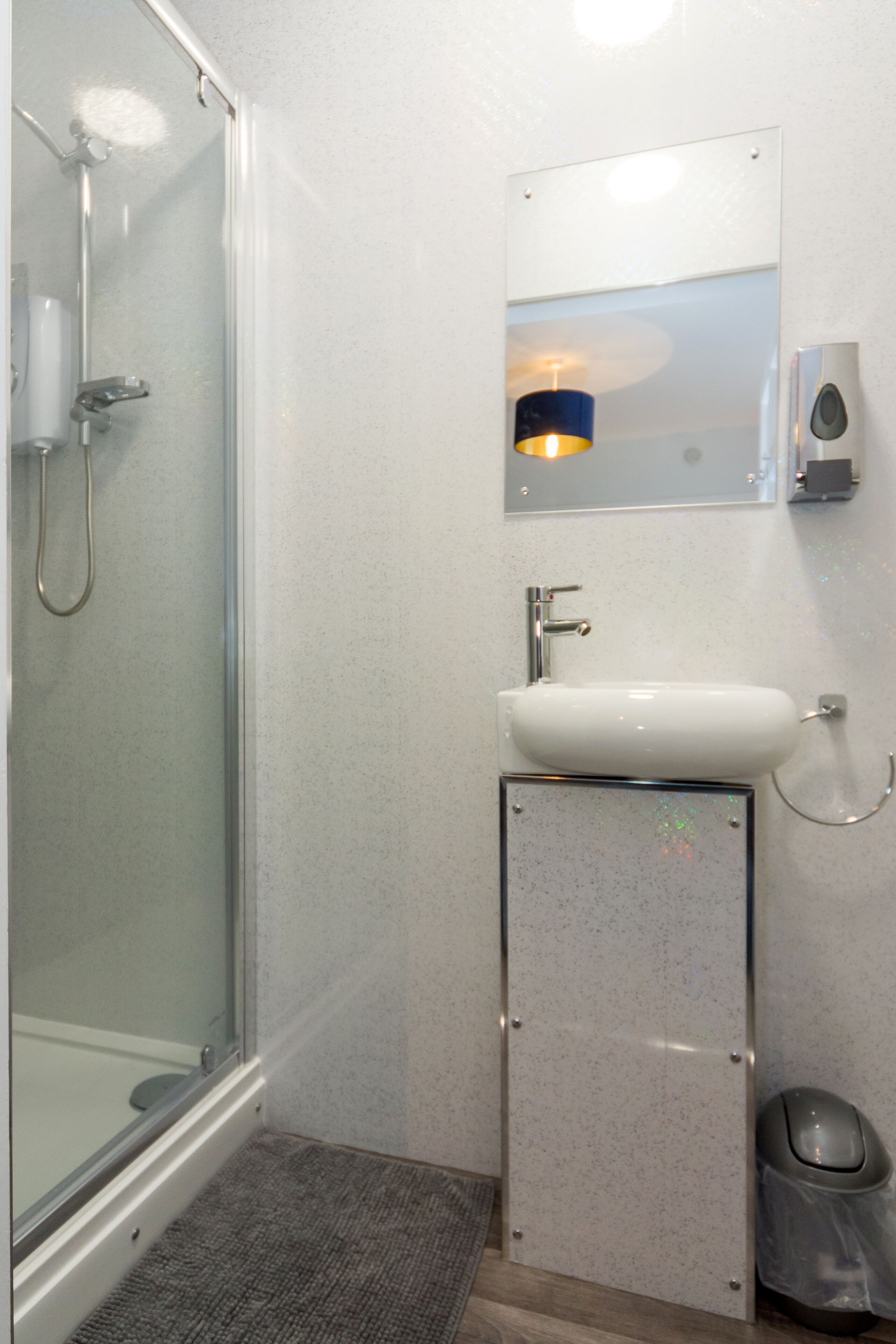 A bathroom with a glass shower enclosure, a white sink, a mirror, and a trash bin.