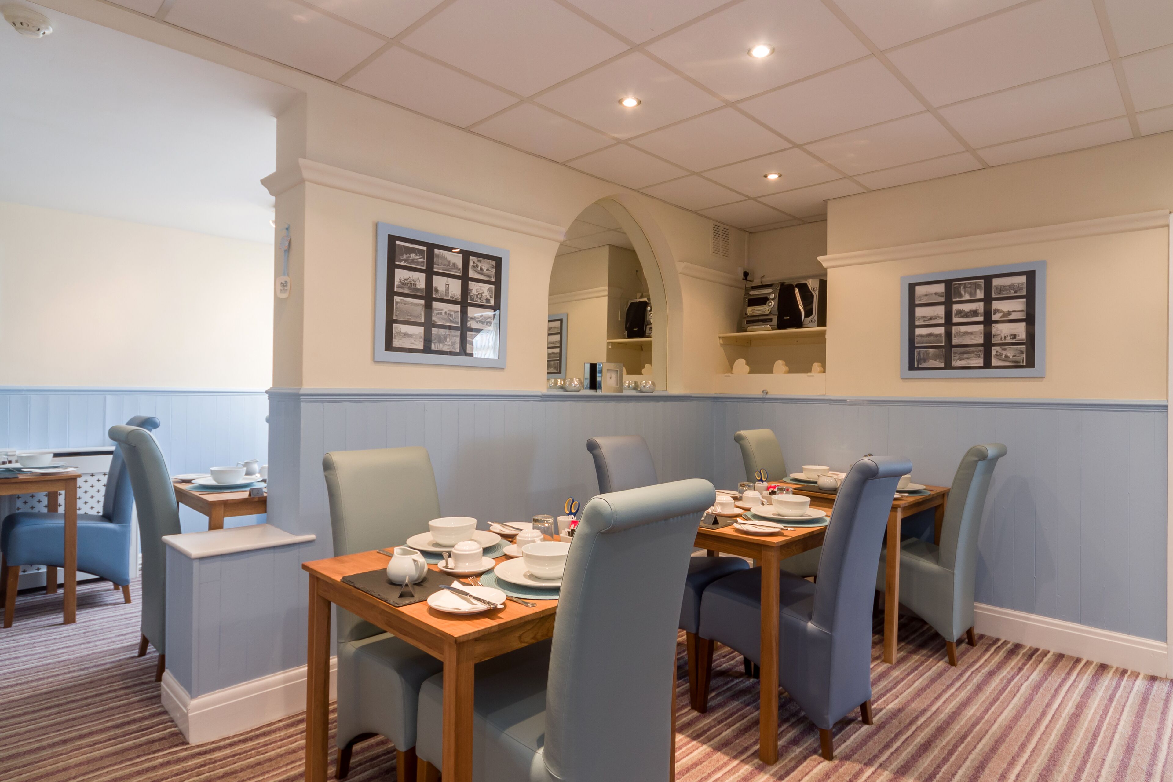 A dining area with tables set for four, light blue upholstered chairs, and a striped carpet.