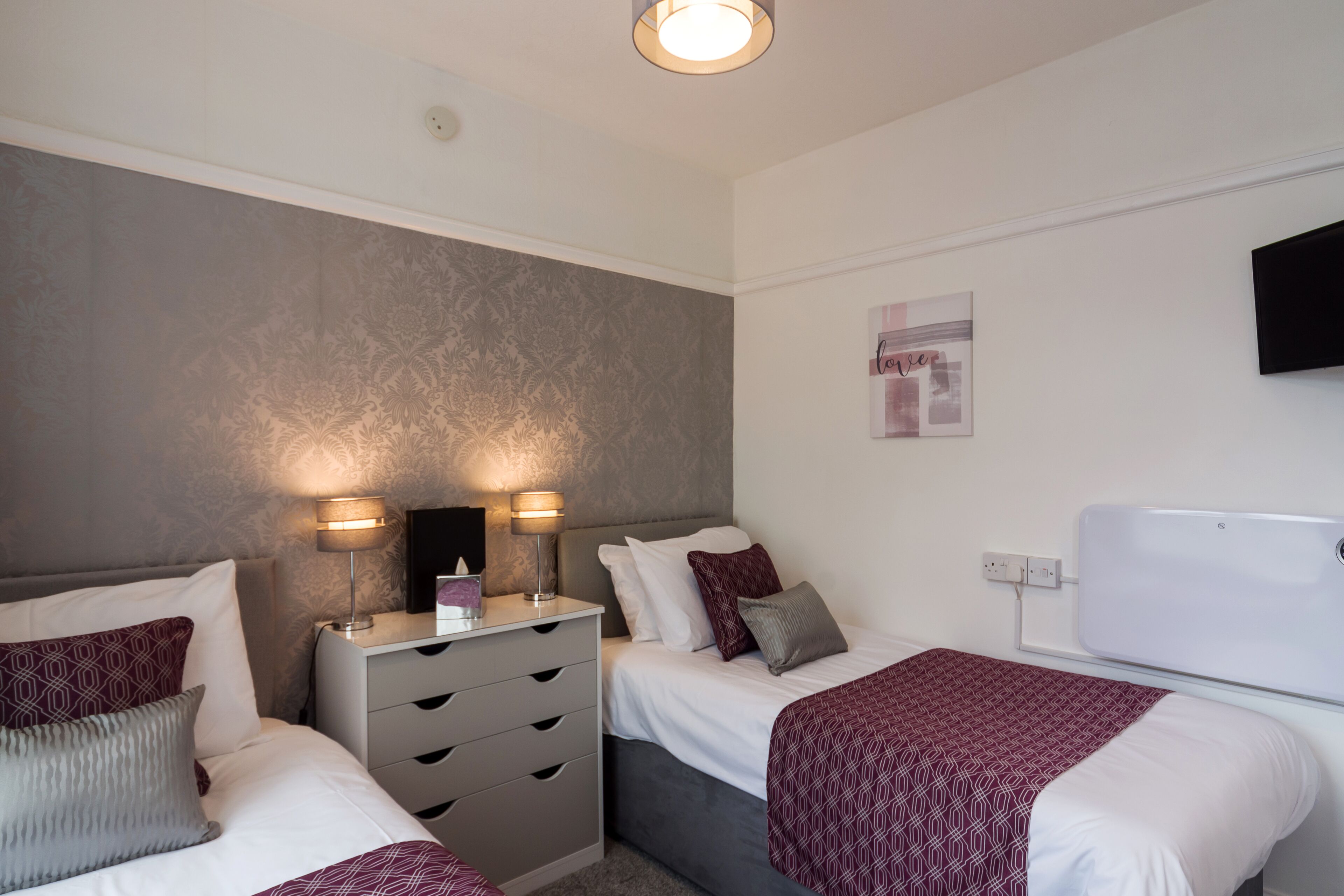Une chambre d’hôtel avec deux lits, une commode, un téléviseur à écran plat et un luminaire fixé au mur.
