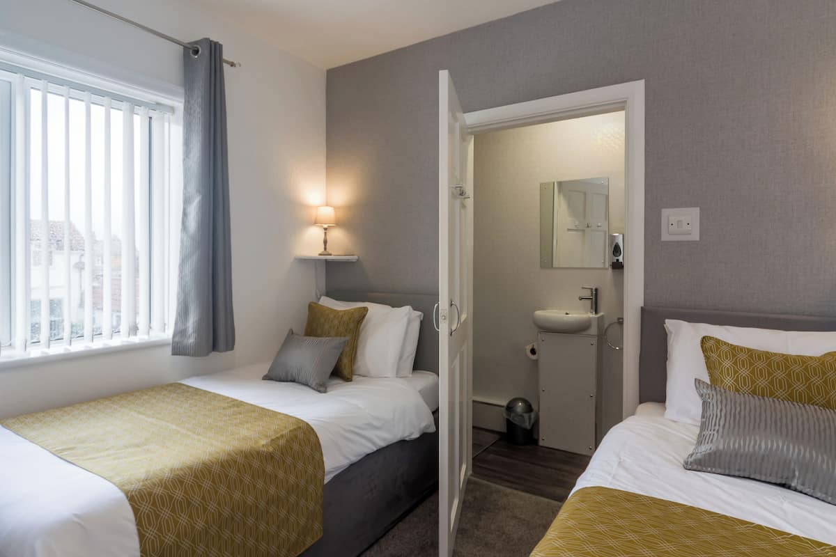 A hotel room with two beds, a window with blinds, a bedside lamp, a bathroom with a sink and mirror, and a door leading to another room.