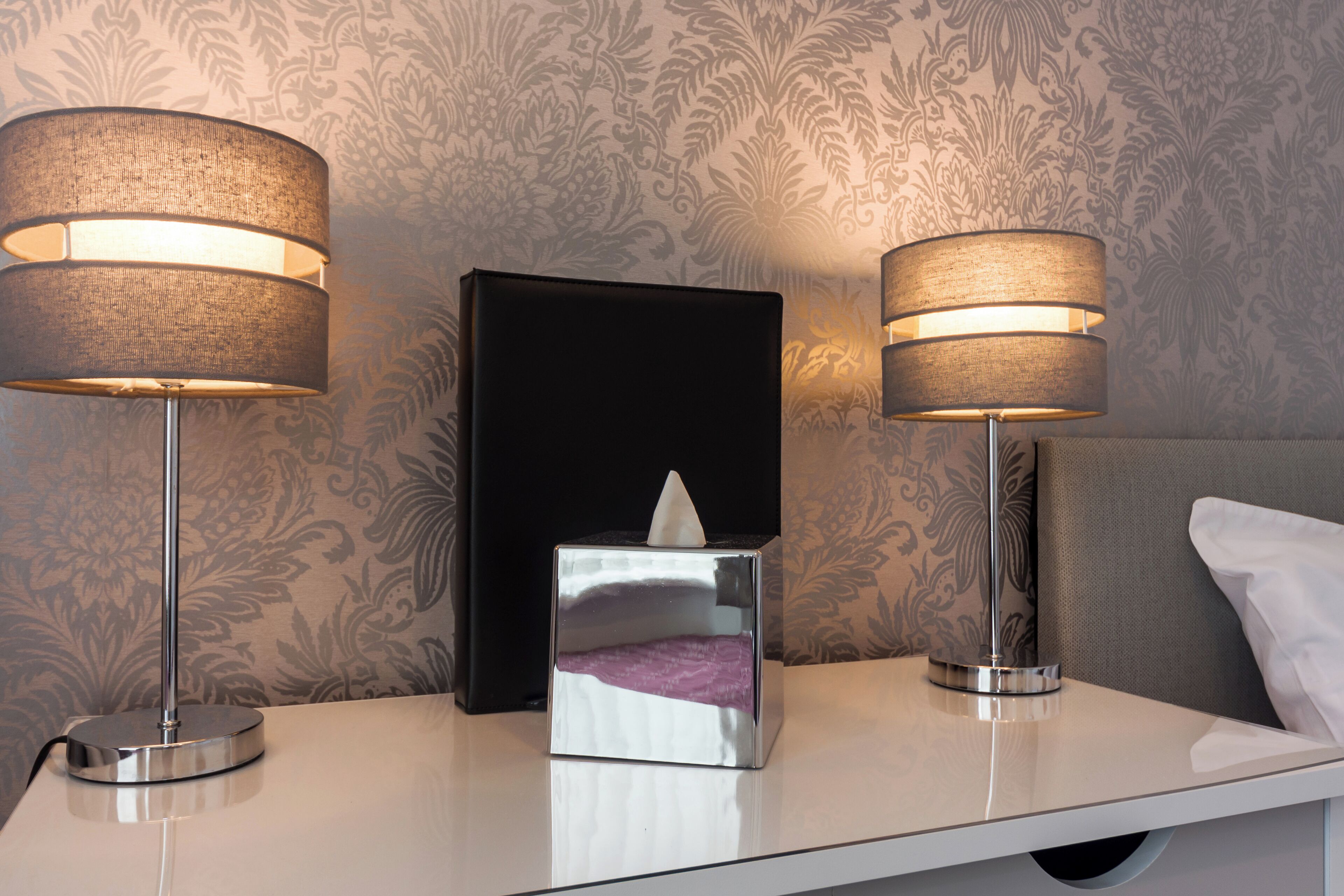 A neatly arranged hotel room with two bedside lamps, a tissue box, and a patterned wallpaper.