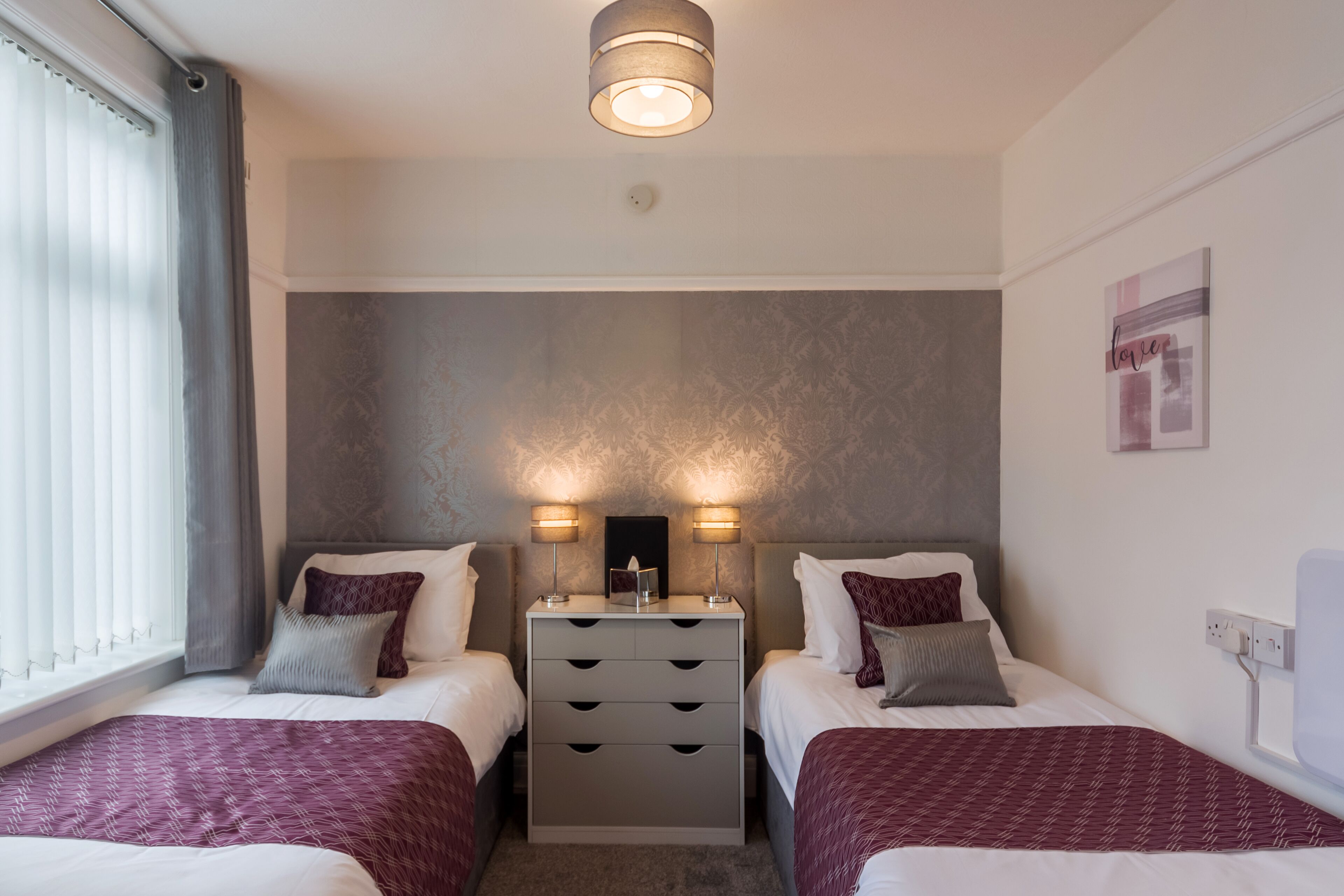 A hotel room with two beds, a nightstand with drawers, a wall-mounted light, and a window with curtains.