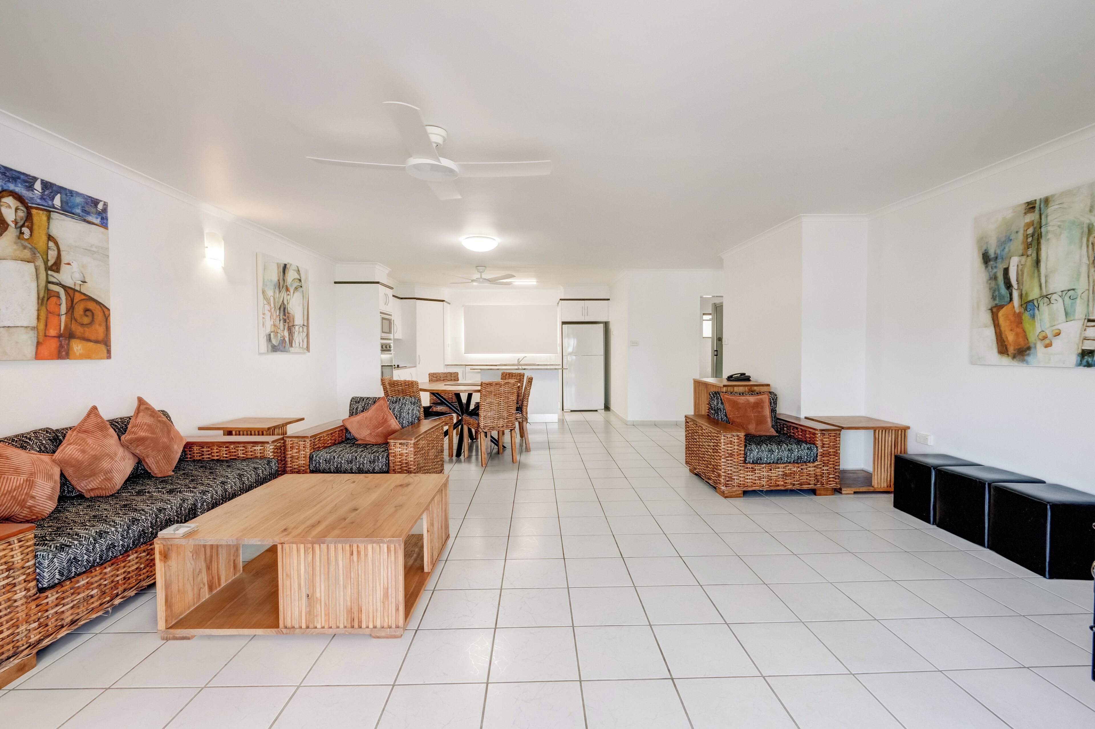 Un vaste espace de vie comprenant un ensemble de canapés, une table basse en bois et une table à manger avec des chaises. Des œuvres d’art sont accrochées aux murs et un ventilateur de plafond est installé.