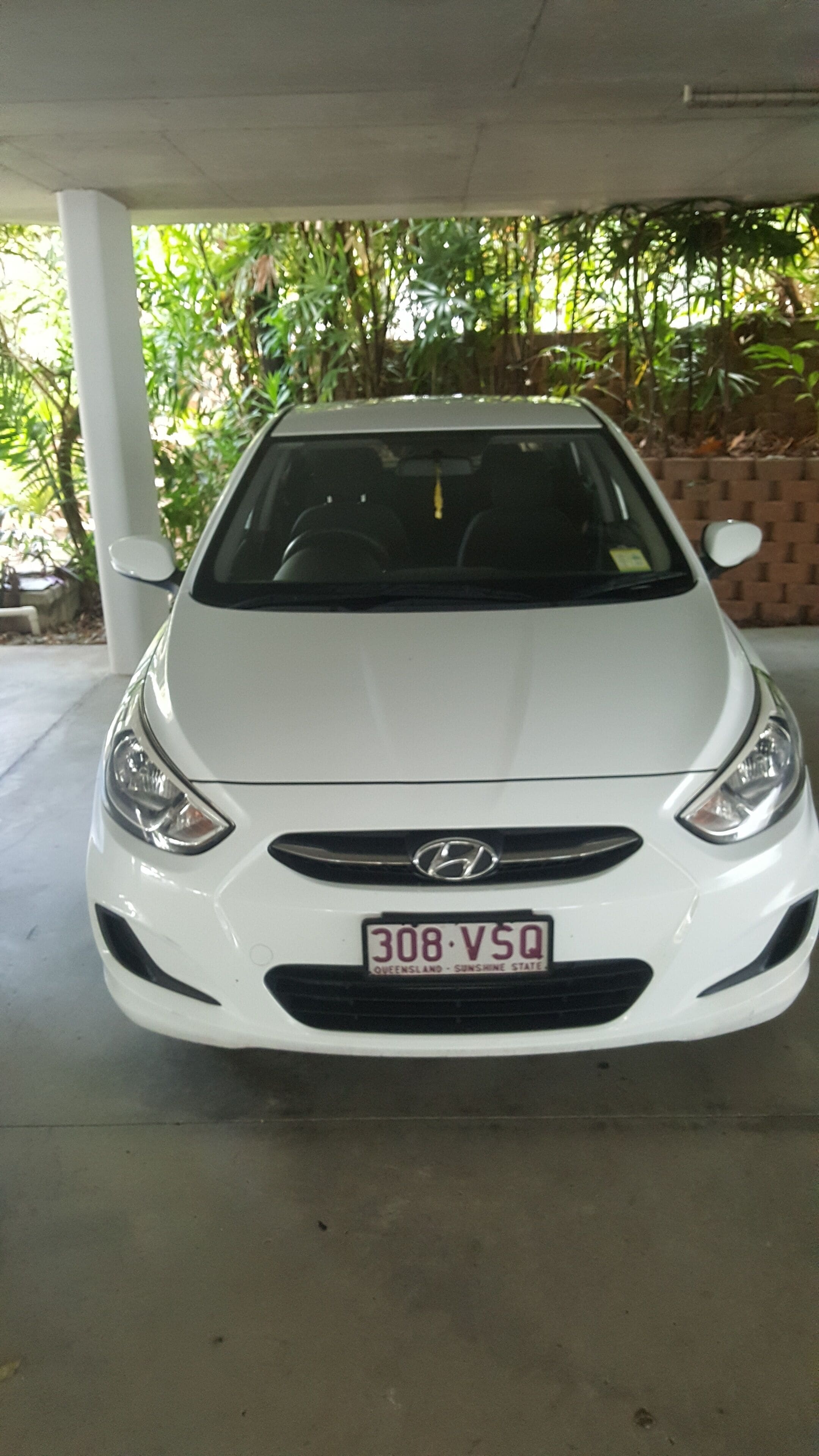 Une voiture Hyundai blanche est garée sous un abri, avec un mur de briques et de la verdure en arrière-plan.