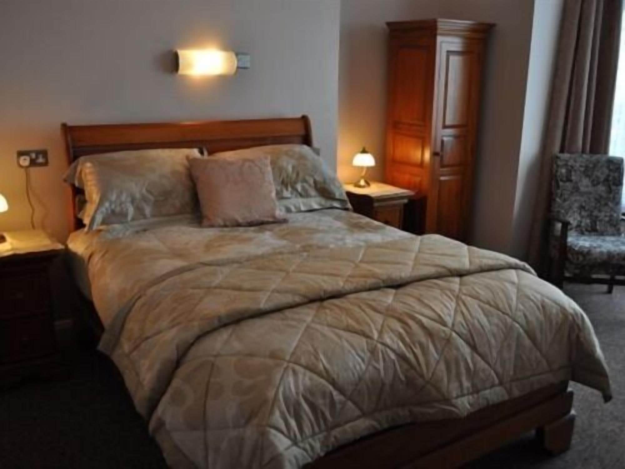Habitación de hotel con una cama grande, dos mesitas de noche con lámparas, un armario de madera y un sillón estampado.