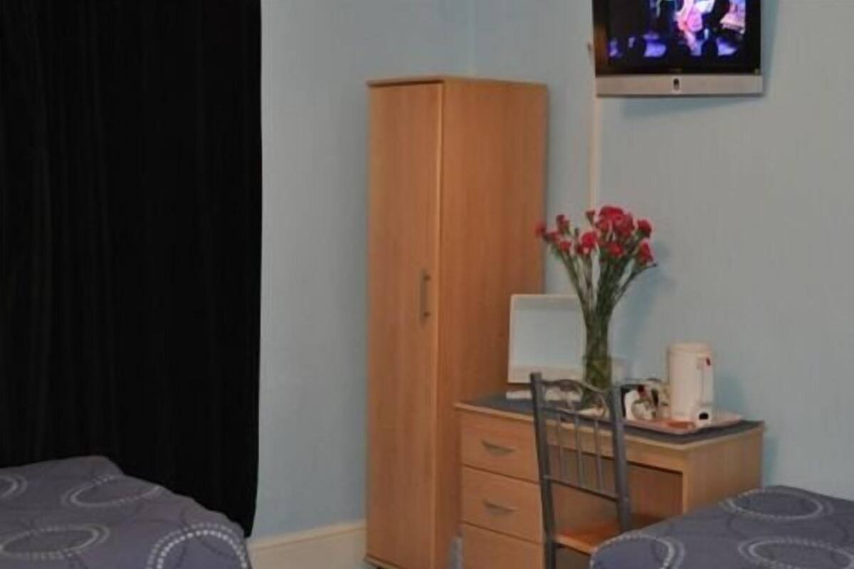 A hotel room with two beds, a wooden wardrobe, a desk with a lamp, a vase with flowers, and a television on the wall.