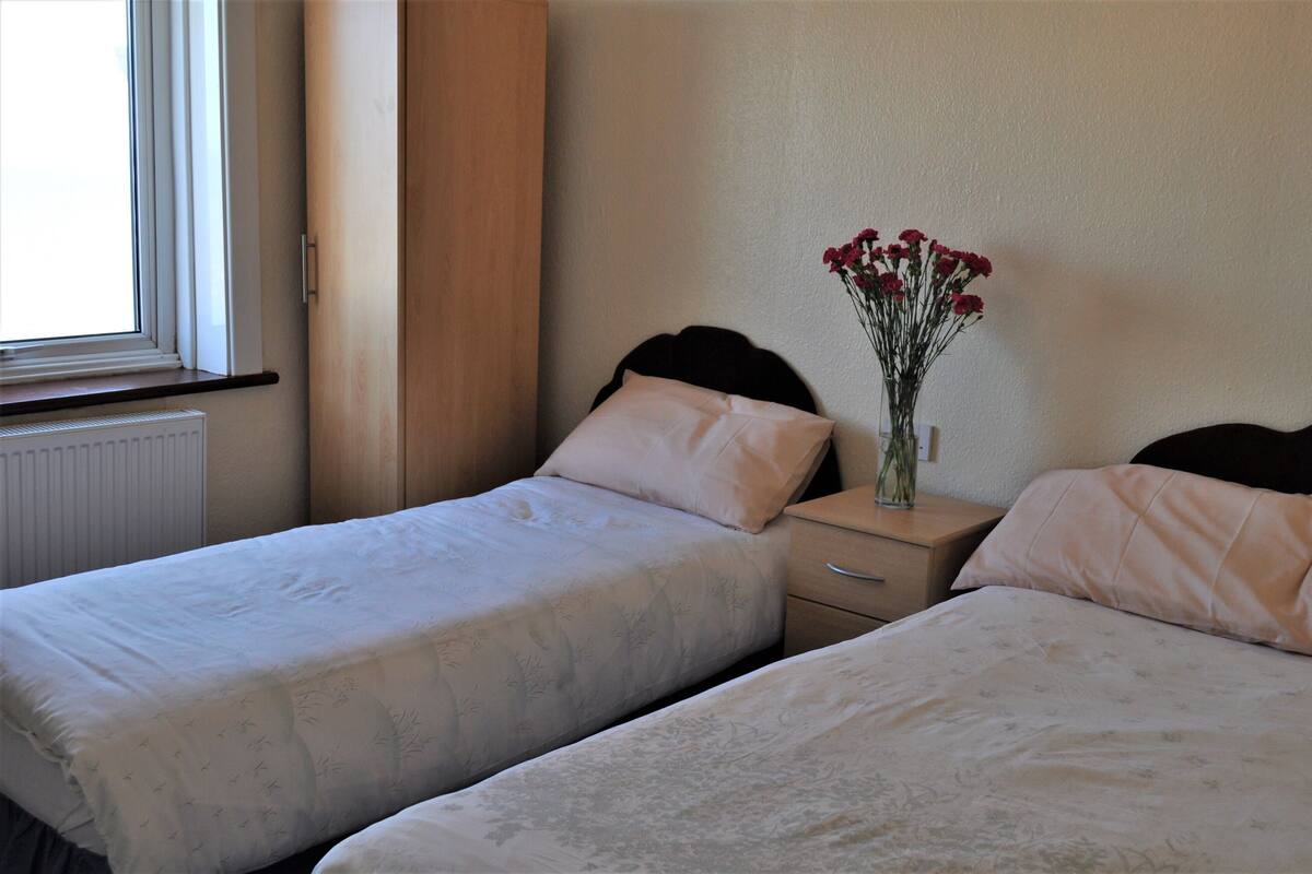 A bedroom with two single beds, a nightstand with a vase of flowers, and a window with curtains.