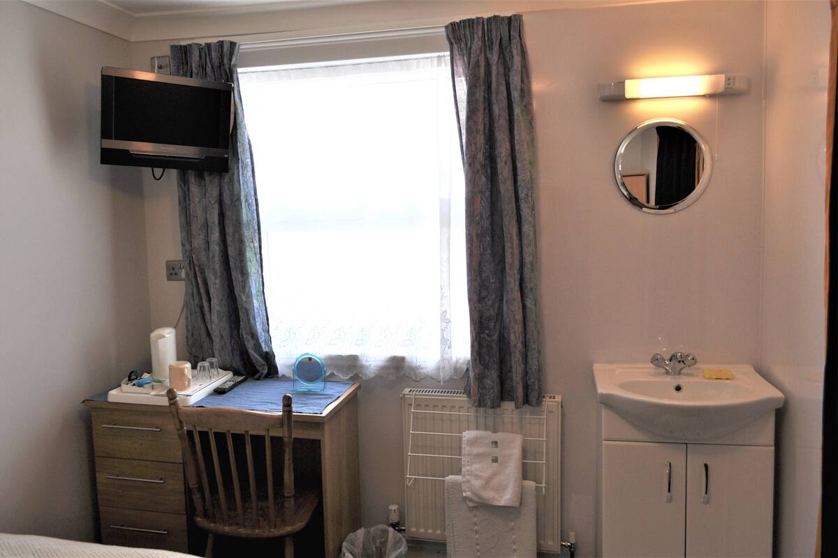 A hotel room with a desk, chair, TV, window with curtains, and a bathroom with a sink and mirror.