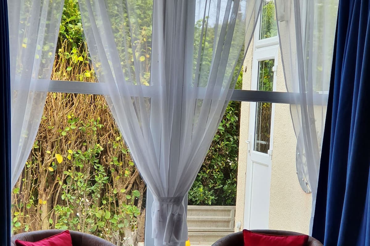A room with two armchairs and a bed, a window with sheer curtains, and a view of greenery outside.