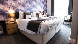 A neatly made bed with a dark blanket, a bedside table with a lamp, and a chair with a cushion. - Grosvenor House (Torquay)