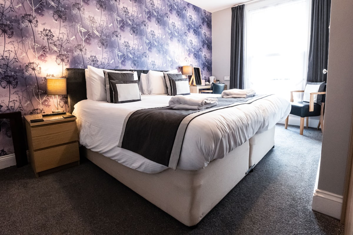 A neatly made bed with a dark blanket, a bedside table with a lamp, and a chair with a cushion.